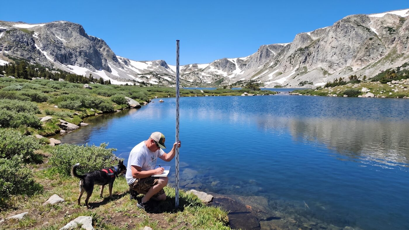 Troy Puls kneeling with a survey rod beside an alpine Snowy Range lake with his service dog